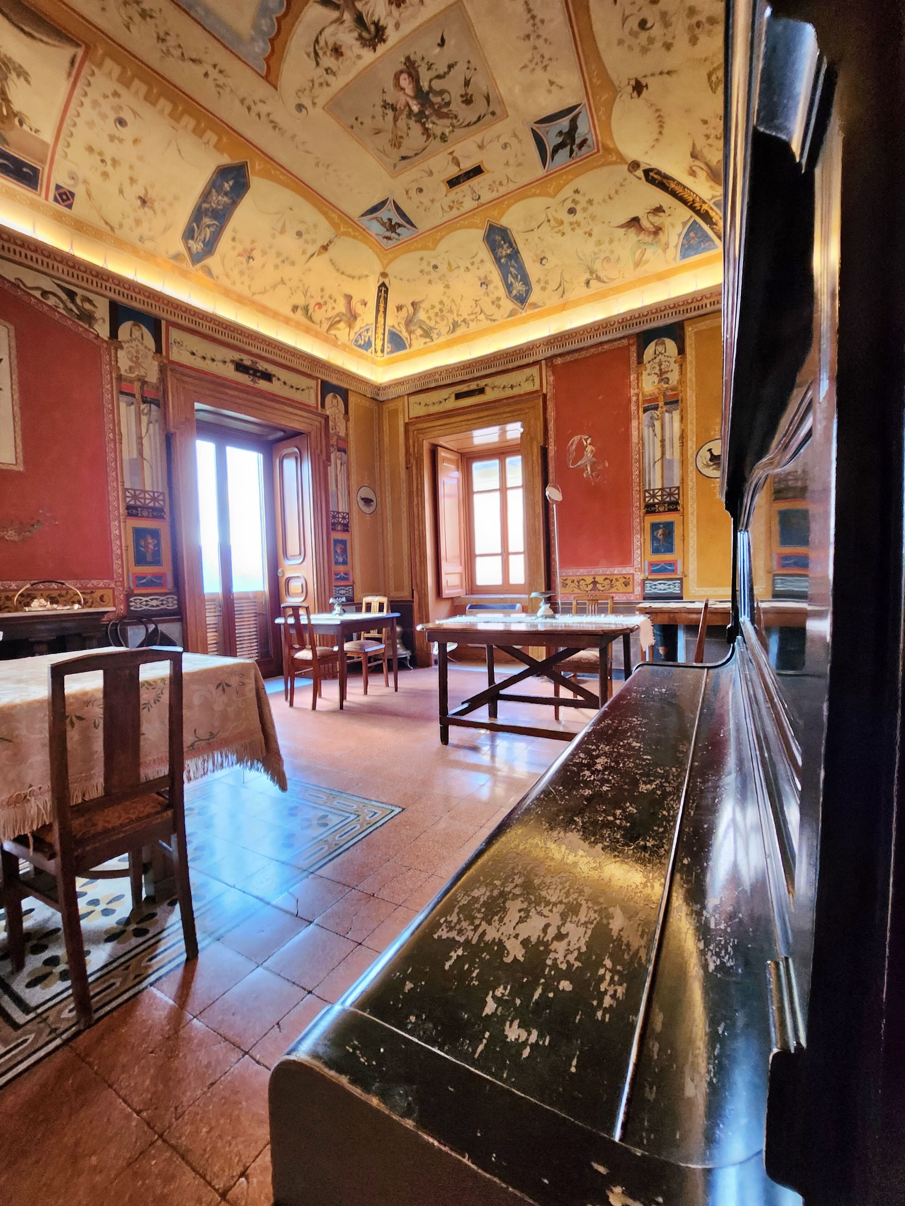 A room with a large wooden bench, a dining table, and chairs. There is a large window with curtains, and the walls are decorated with floral patterns.