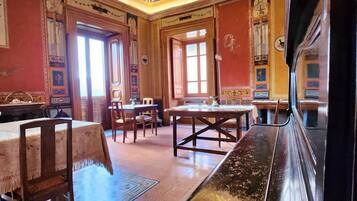 A room with a large wooden bench, a dining table, and chairs. There is a large window with curtains, and the walls are decorated with floral patterns.