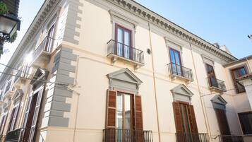 Un edificio a più piani con elementi architettonici classici, tra cui finestre simmetriche con persiane, un balcone con ringhiera in ferro battuto e una cornice decorativa.