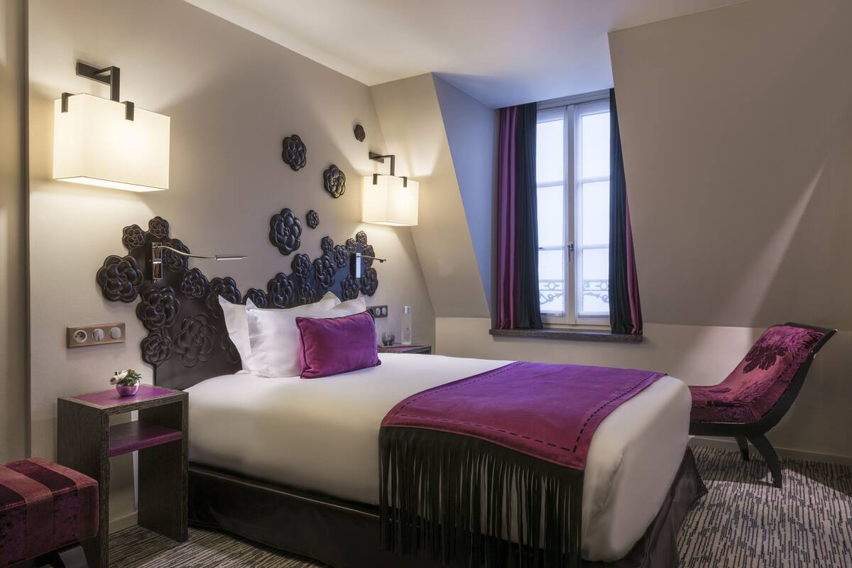 A modern hotel room with a large bed, decorative wall art, two bedside lamps, a window with curtains, and a chair.