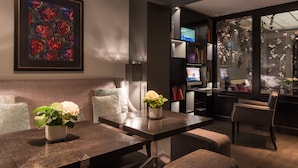 A modern living room with a dark wood coffee table, a white vase with flowers, a large framed artwork, and a bookshelf with books and a television. - Hôtel Les Jardins de La Villa (Paris)