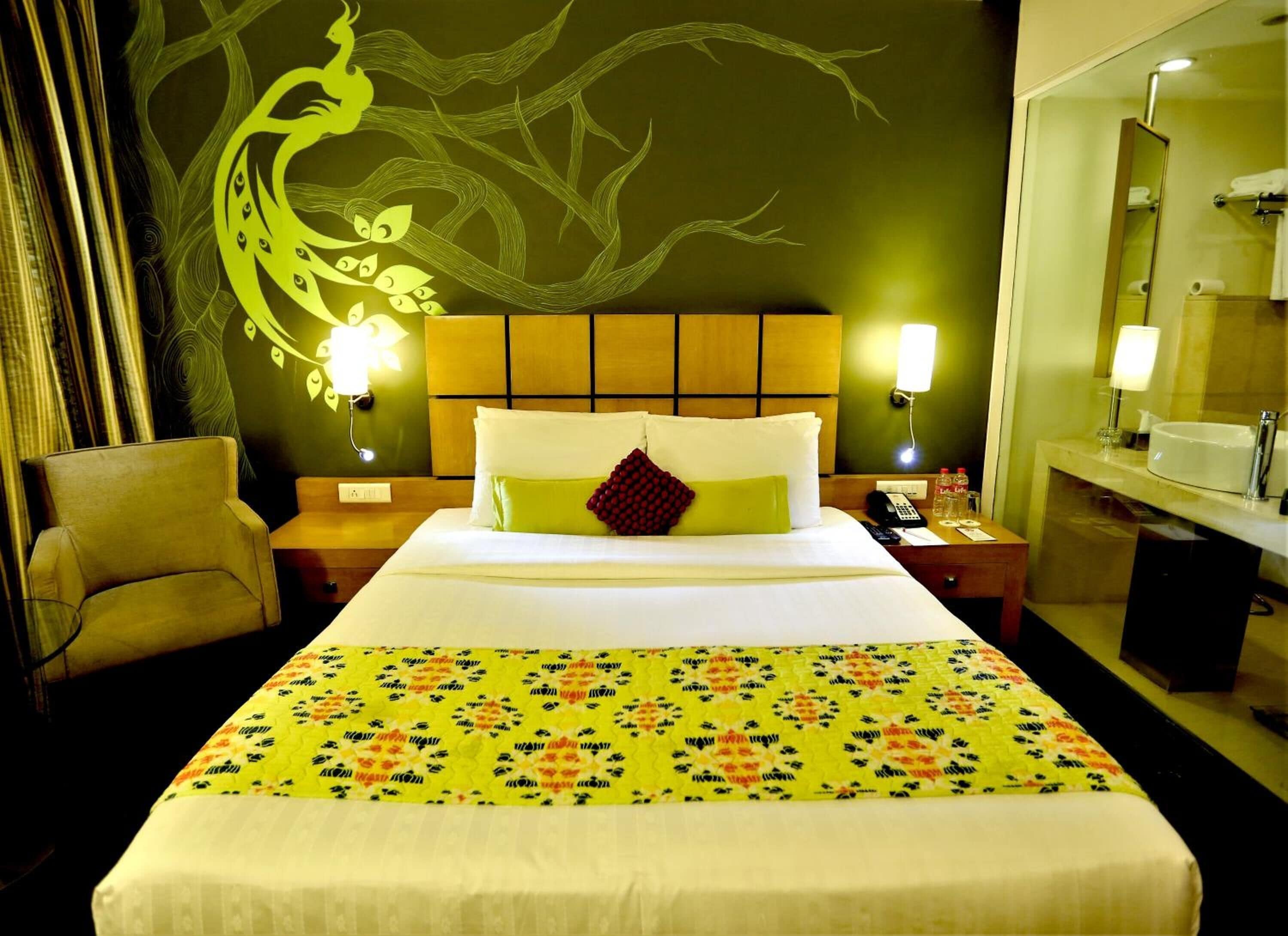 A hotel room with a large bed, a chair, a nightstand, a mirror, and a wall with a tree design.