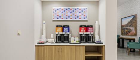 A coffee station with labeled dispensers for light, dark, hot, and decaf coffee, a painting on the wall, and a seating area with a table and chairs.