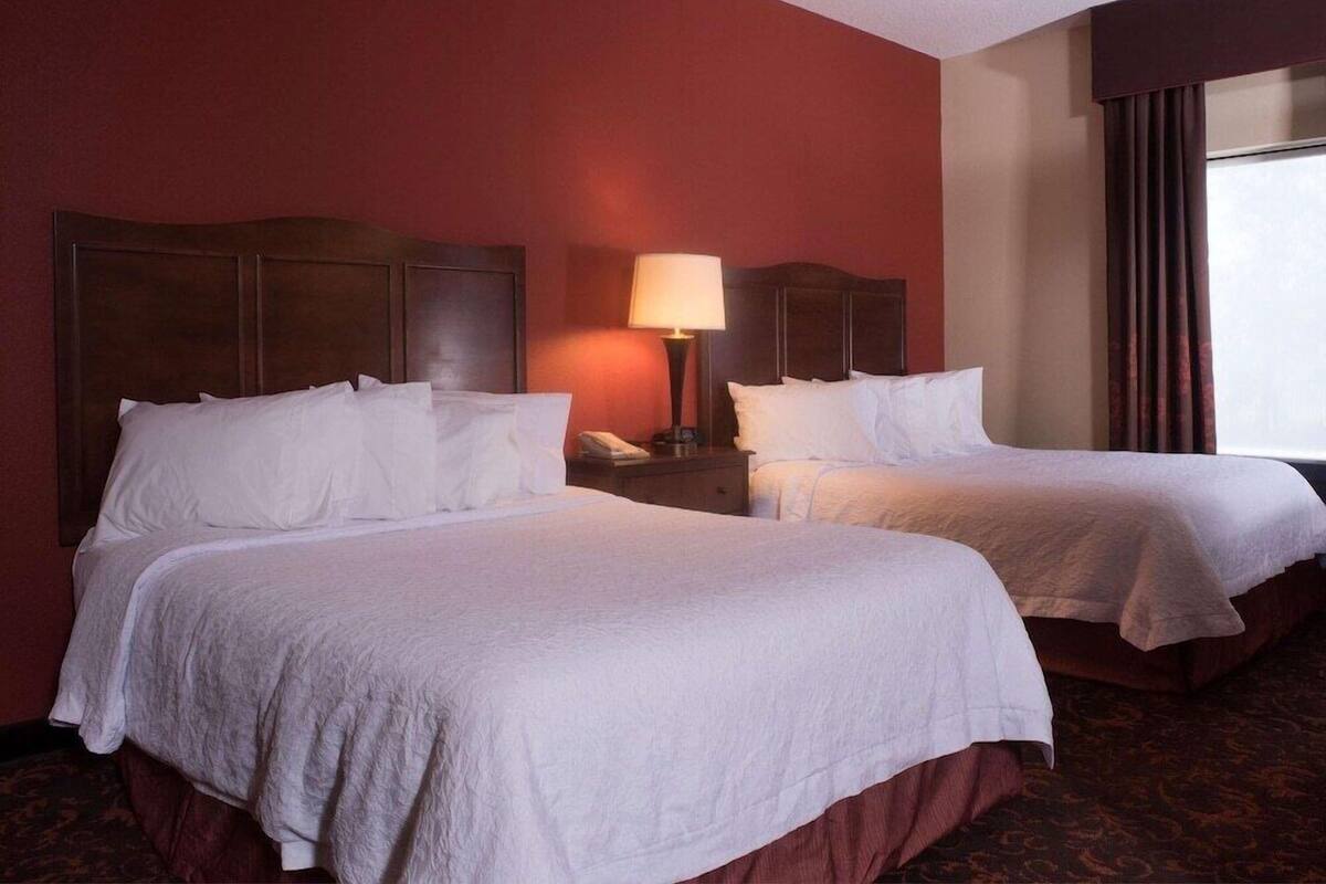 A hotel room with two beds, a nightstand with a lamp, and a window with curtains.