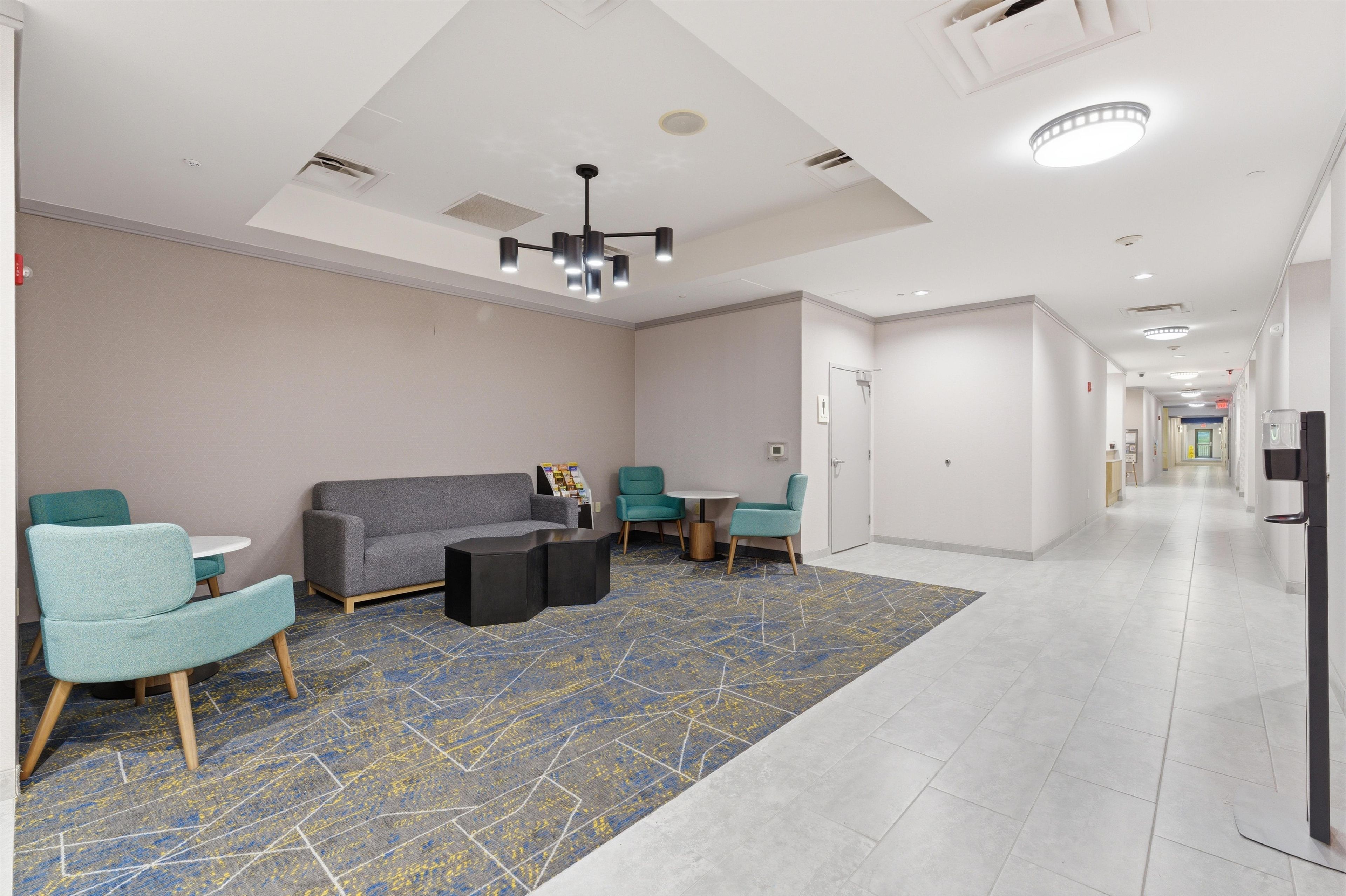 A modern hotel lobby with a sofa, chairs, a coffee table, a patterned rug, and a hallway leading to other rooms.