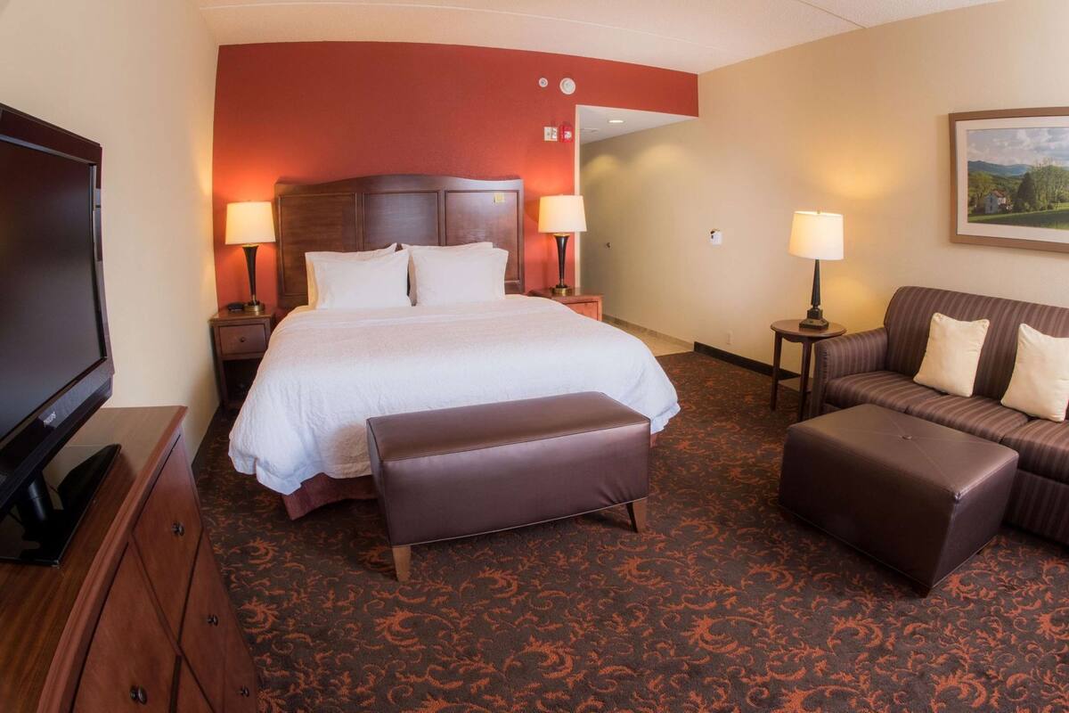 A hotel room with a bed, a TV, a sofa, an ottoman, and a painting on the wall.