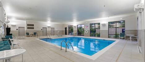 Una amplia piscina interior con agua cristalina y azul, rodeada de suelo de baldosas blancas y sillas verdes.