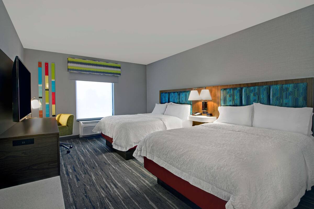 A hotel room with two beds, a TV, a desk, a chair, a window, and a colorful wall decoration.
