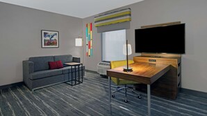 Habitación de hotel con sofá gris, escritorio de madera, televisor de pantalla plana, ventana con vistas y una obra de arte abstracta y colorida en la pared.