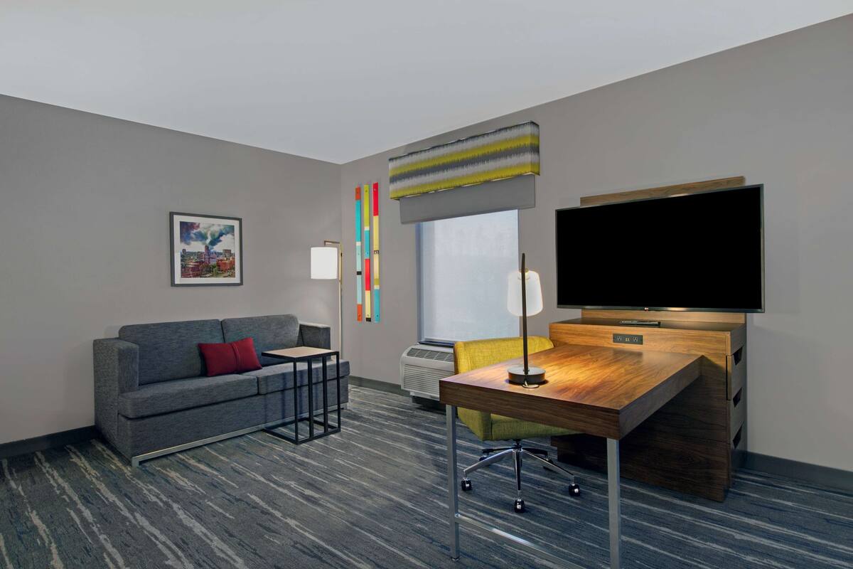 A hotel room with a gray sofa, a wooden desk, a flat-screen TV, a window with a view, and a colorful abstract art piece on the wall.