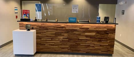 A hotel reception desk with a wooden panel design, a digital payment terminal, and signs indicating social distancing measures.
