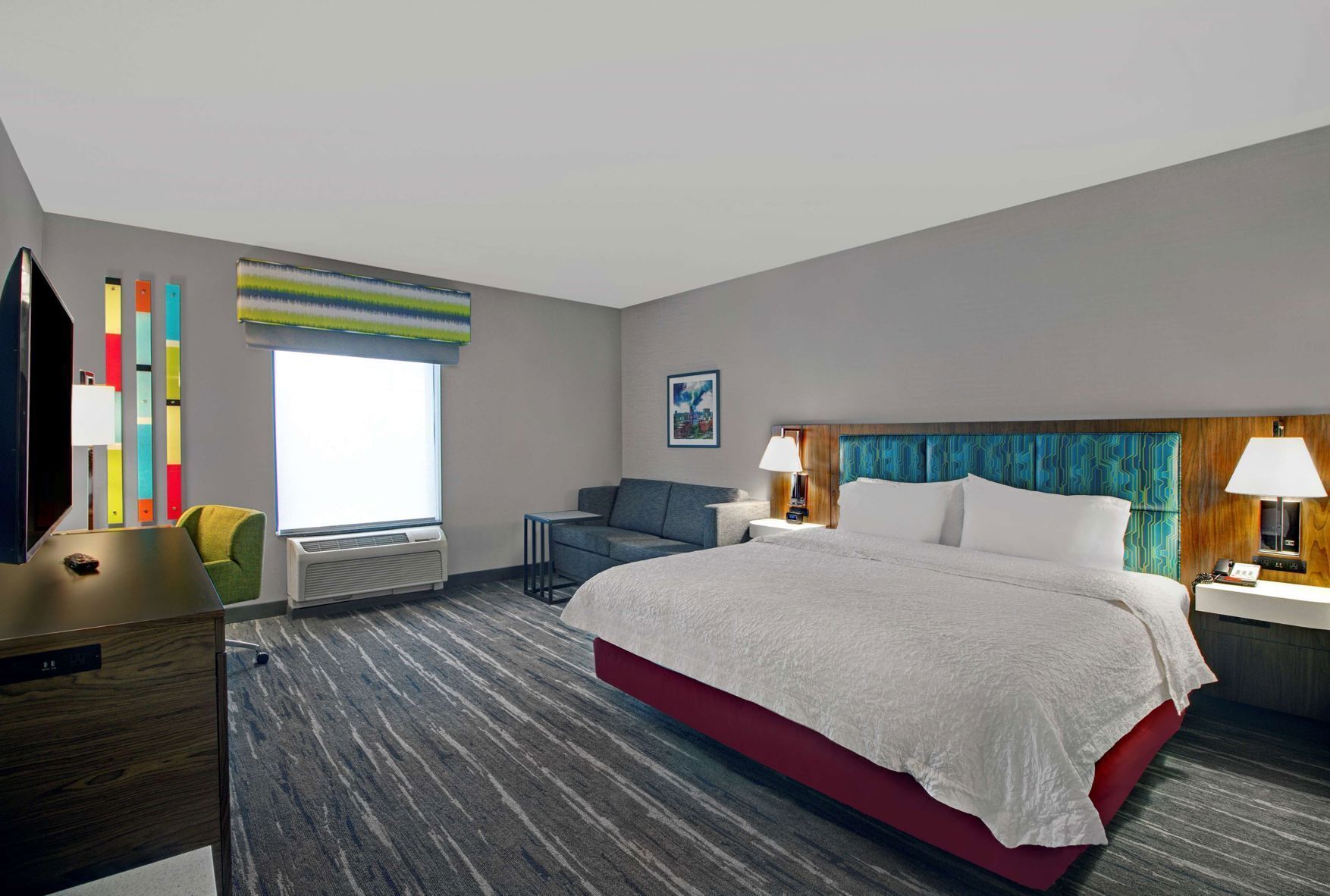A hotel room with a large bed, a desk, a chair, a TV, a window with blinds, and a wall with colorful vertical blinds.