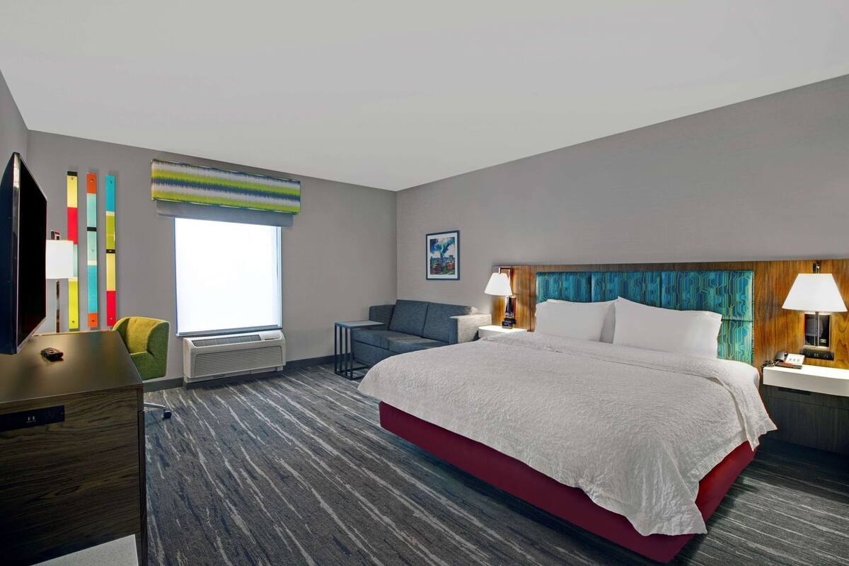Habitación de hotel con una cama grande, un escritorio, una silla, un televisor, una ventana con persianas y una pared con persianas verticales coloridas.
