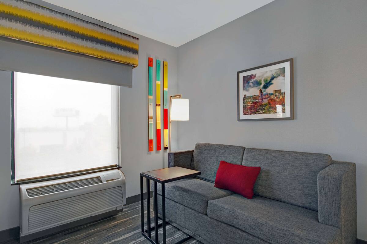 A modern hotel room with a grey sofa, a small table, a radiator, and colorful abstract art on the wall.