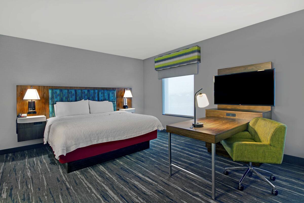 A hotel room with a bed, bedside tables, a desk, a chair, a TV, and a window.