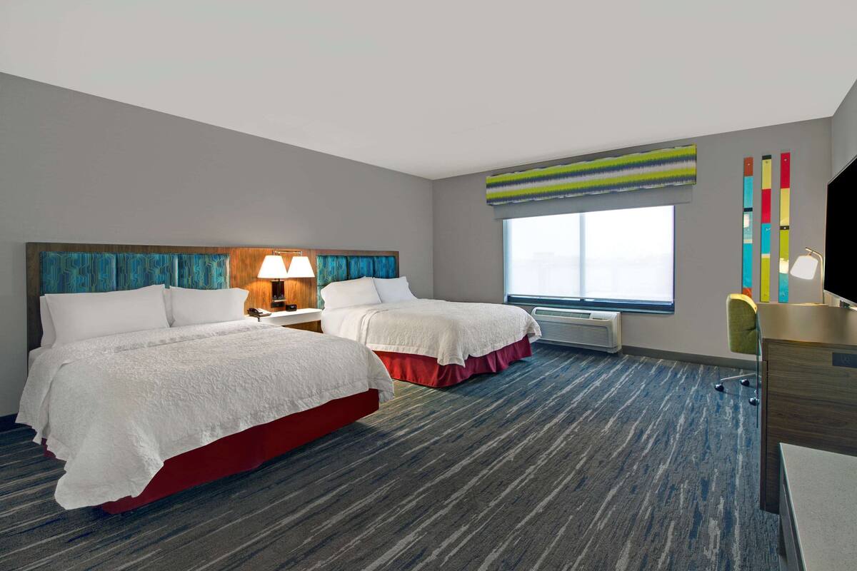 A hotel room with two beds, a desk, a chair, a lamp, a TV, and a window with a view.