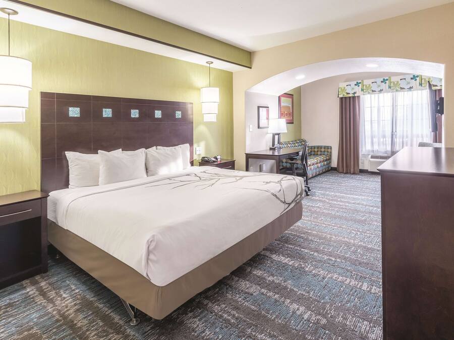 Habitación de hotel con una cama grande, un escritorio con silla, una mesita pequeña y un sofá. Se aprecia una ventana con cortinas y vistas al exterior.