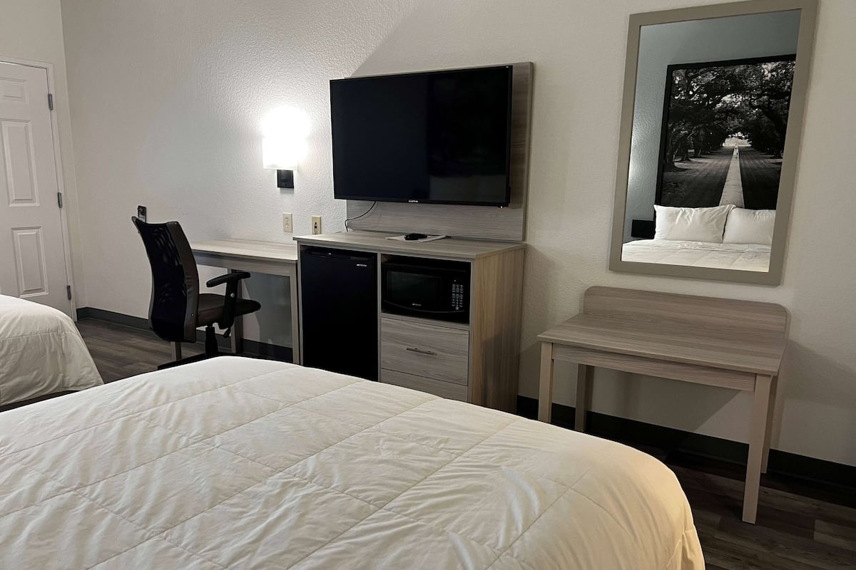 Quarto de hotel com cama, escrivaninha com televisão, cadeira, micro-ondas e uma mesinha.