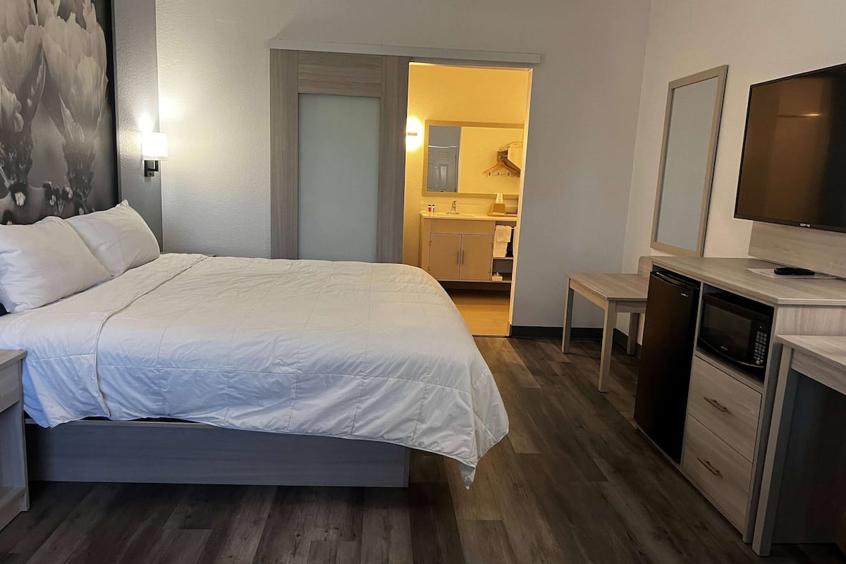 Quarto de hotel com uma cama grande, uma mesinha, uma televisão e uma cozinha compacta.