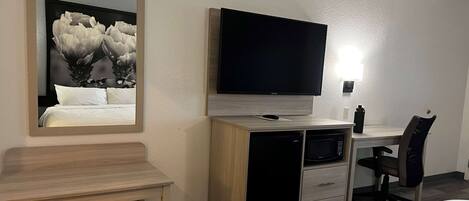 A hotel room with a bed, a desk with a flat-screen TV, a microwave, a refrigerator, a chair, and a bedside table.