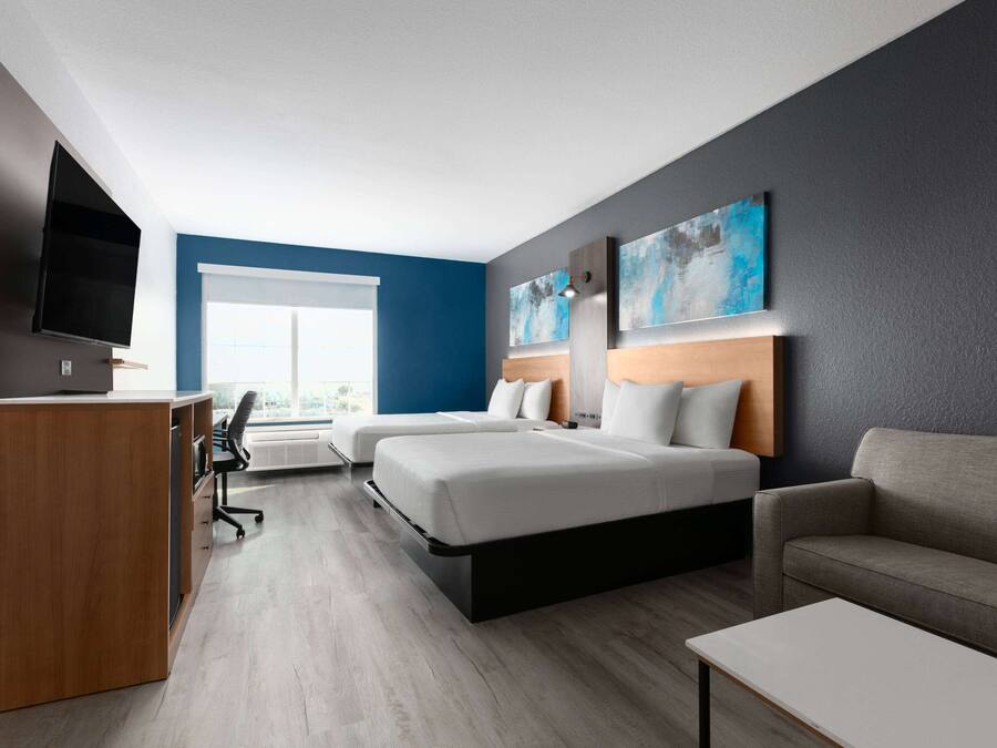 A modern hotel room with a desk, two beds, a sofa, and a large window.