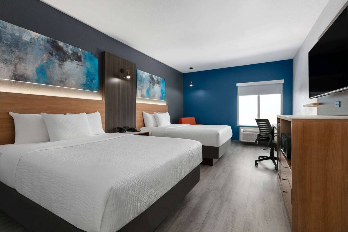 Quarto de hotel moderno com duas camas, uma mesa, uma cadeira, televisão de tela plana e uma grande pintura abstrata na parede.