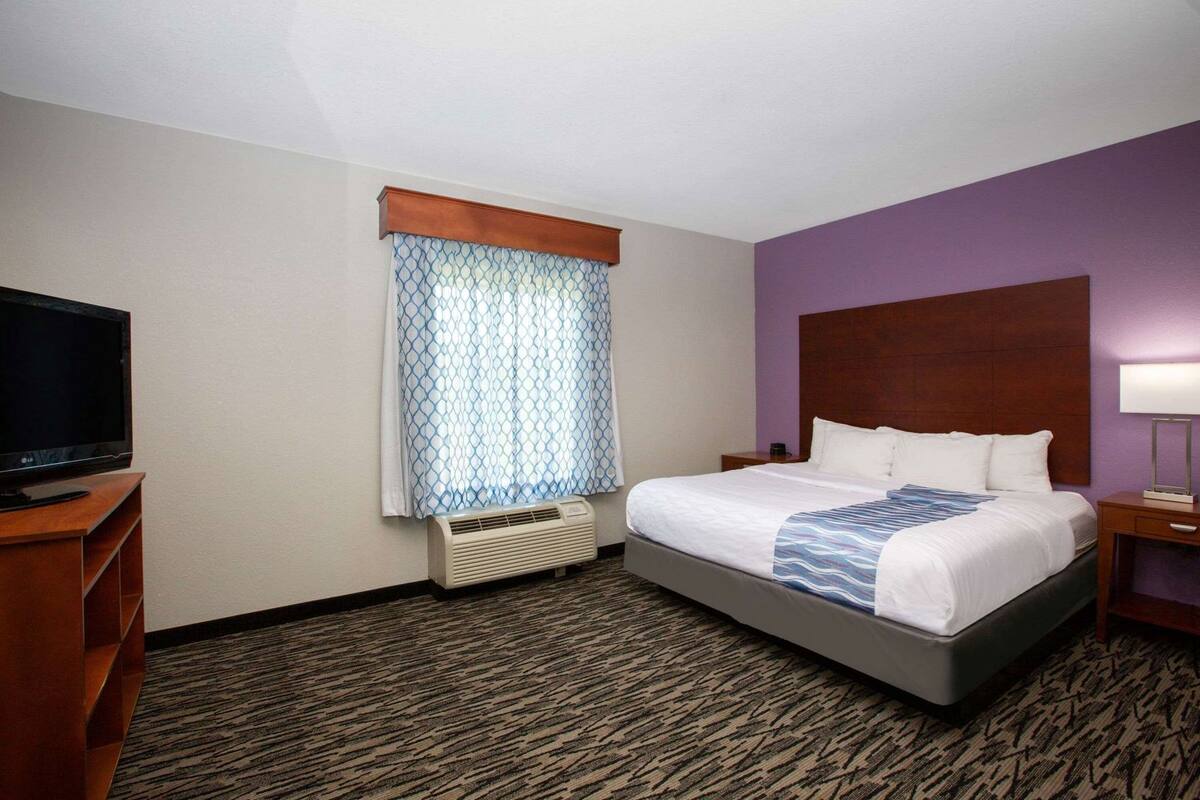 A hotel room with a bed, a television, an air conditioning unit, and a window with patterned curtains.