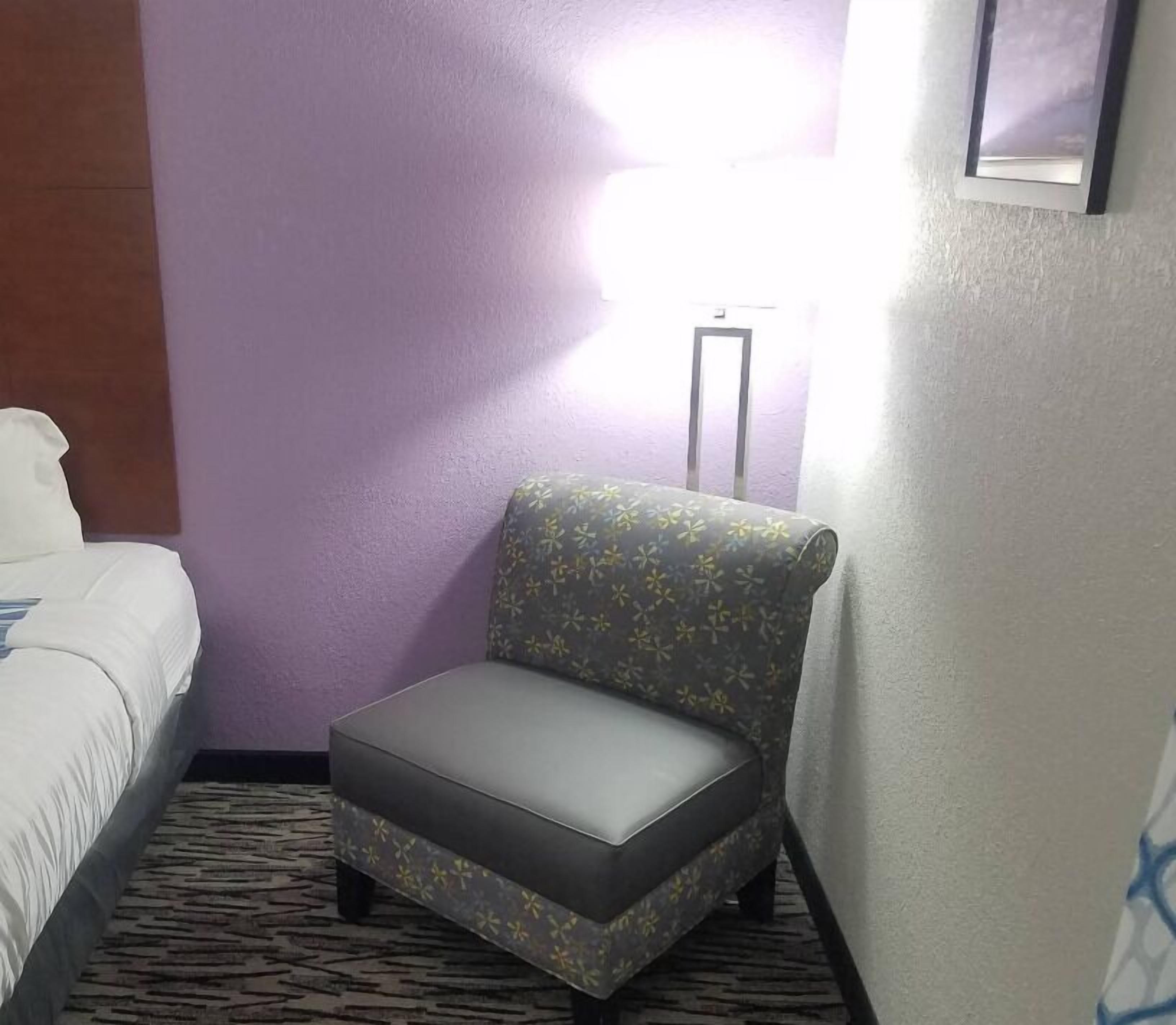 A hotel room with a bed, a gray armchair with a floral pattern, and a standing lamp.