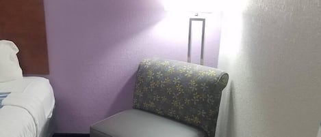 A hotel room with a bed, a gray armchair with a floral pattern, and a standing lamp.