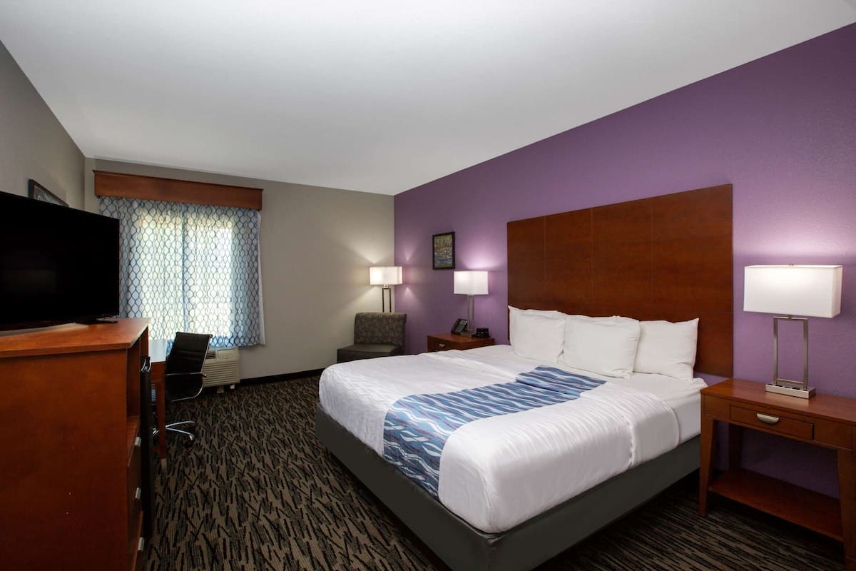 A hotel room with a large bed, a desk with a flat-screen TV, a chair, two bedside lamps, and a window with patterned curtains.