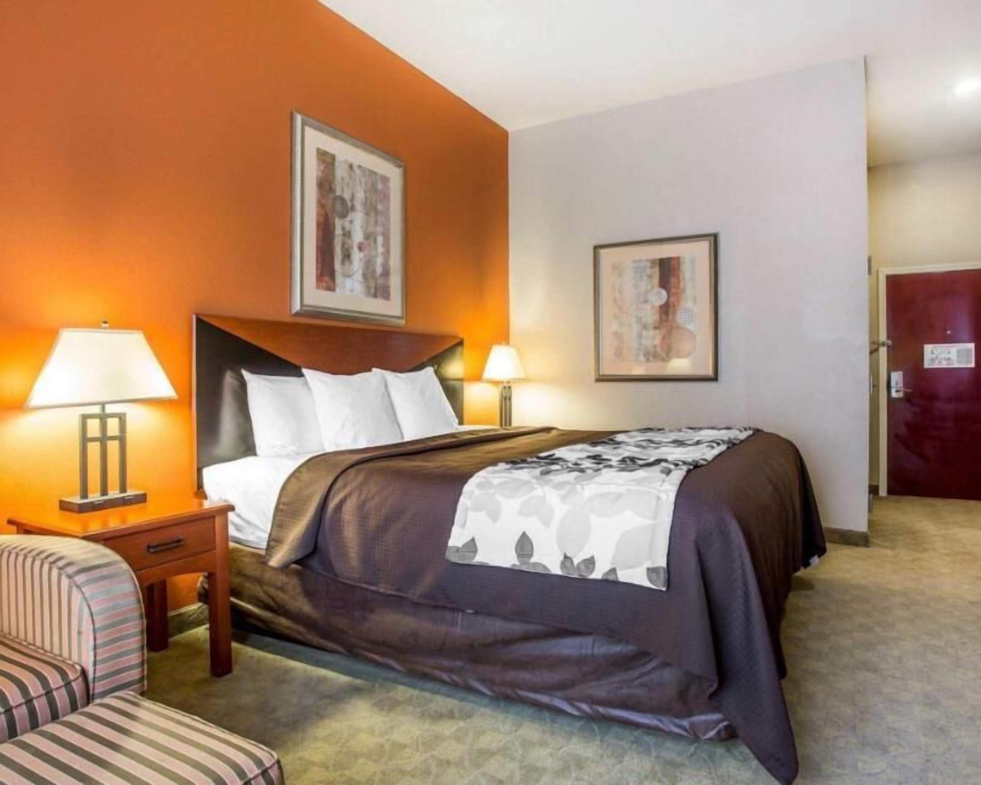 Quarto de hotel com cama, duas luminárias de cabeceira, uma cadeira e duas quadros pendurados na parede.