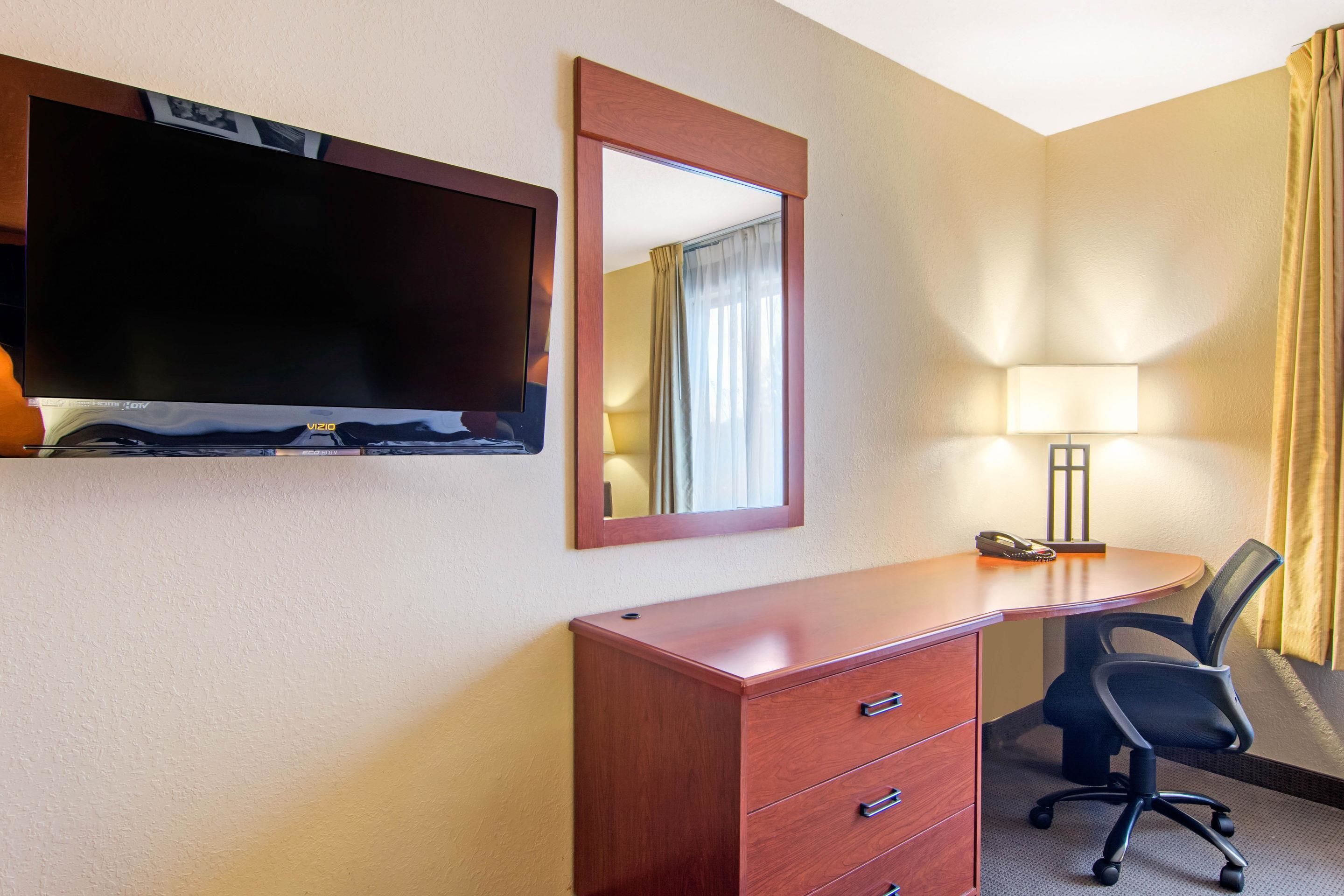 A hotel room with a wooden desk, a TV, a mirror, a lamp, and a chair.