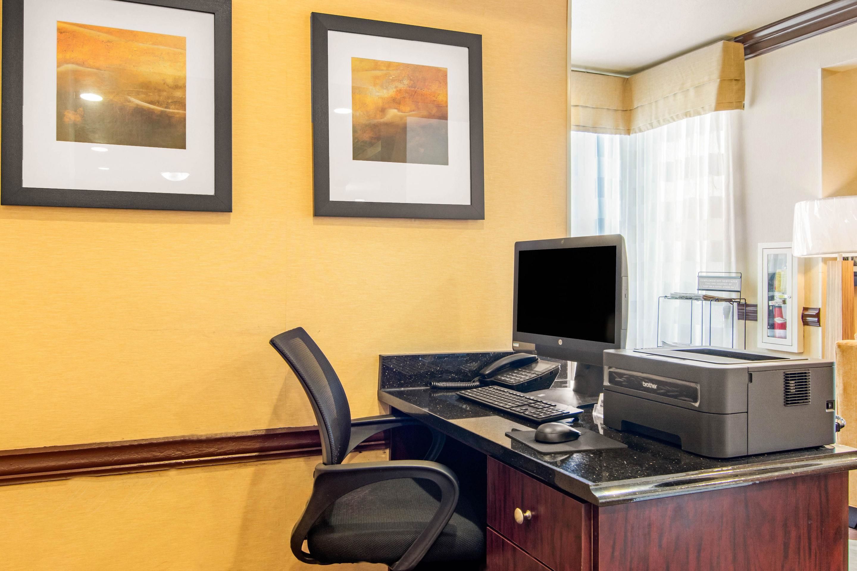 A hotel room with a desk, computer, printer, and framed artwork on the wall.