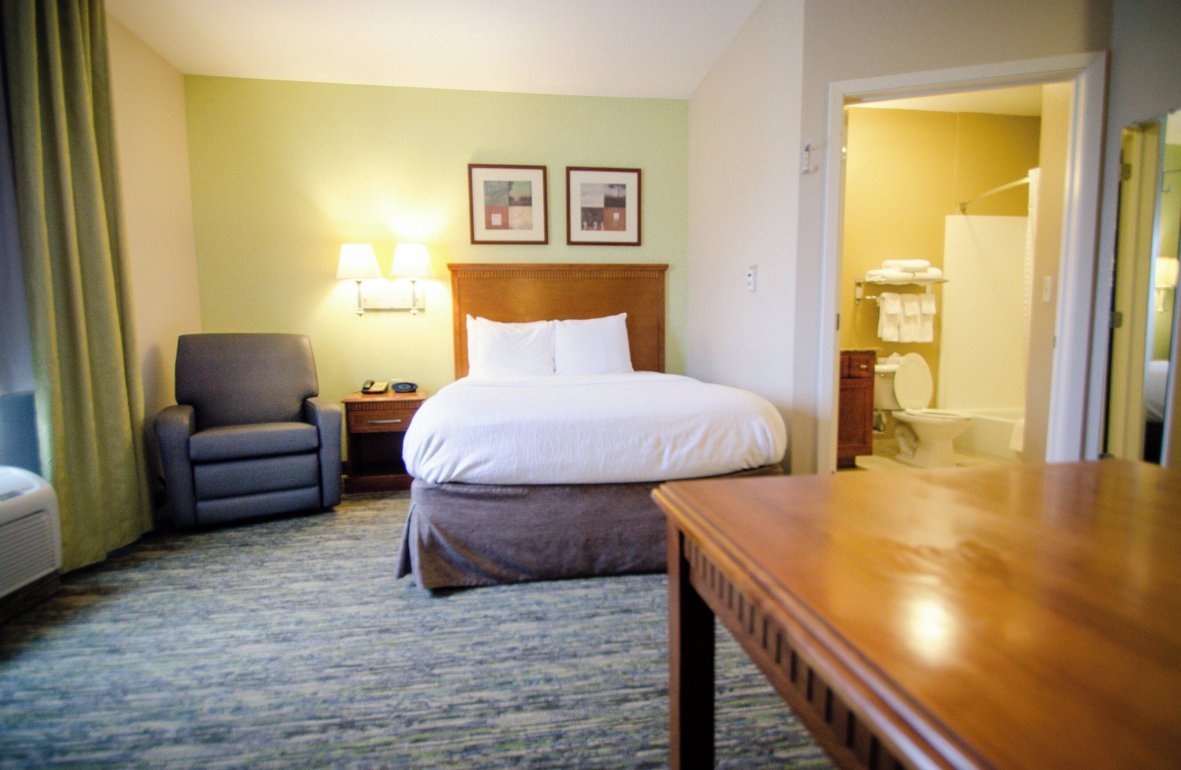 Une chambre d’hôtel avec un lit, une chaise, une table de chevet, une lampe et une salle de bain visible à travers une porte ouverte.