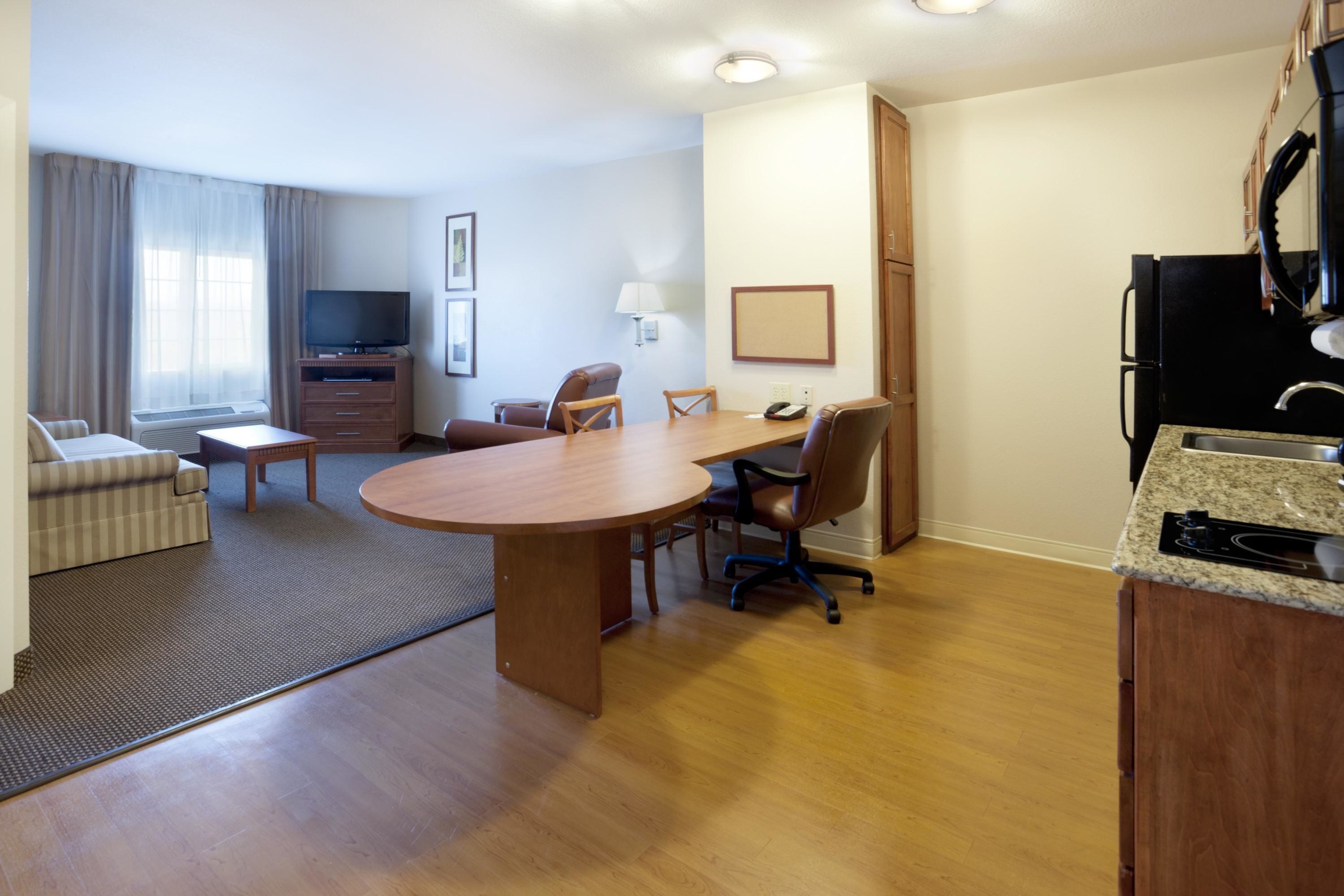 A hotel room with a dining table, chairs, a sofa, a TV, a lamp, a refrigerator, and a kitchen counter.