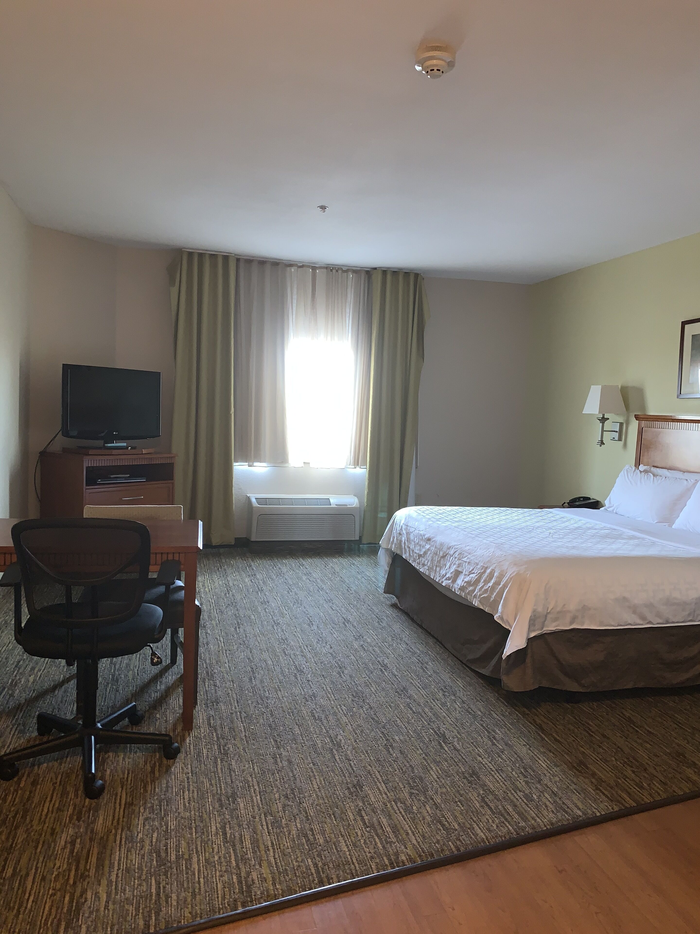 A hotel room with a bed, a desk with a television, a chair, a window with curtains, and a lamp.