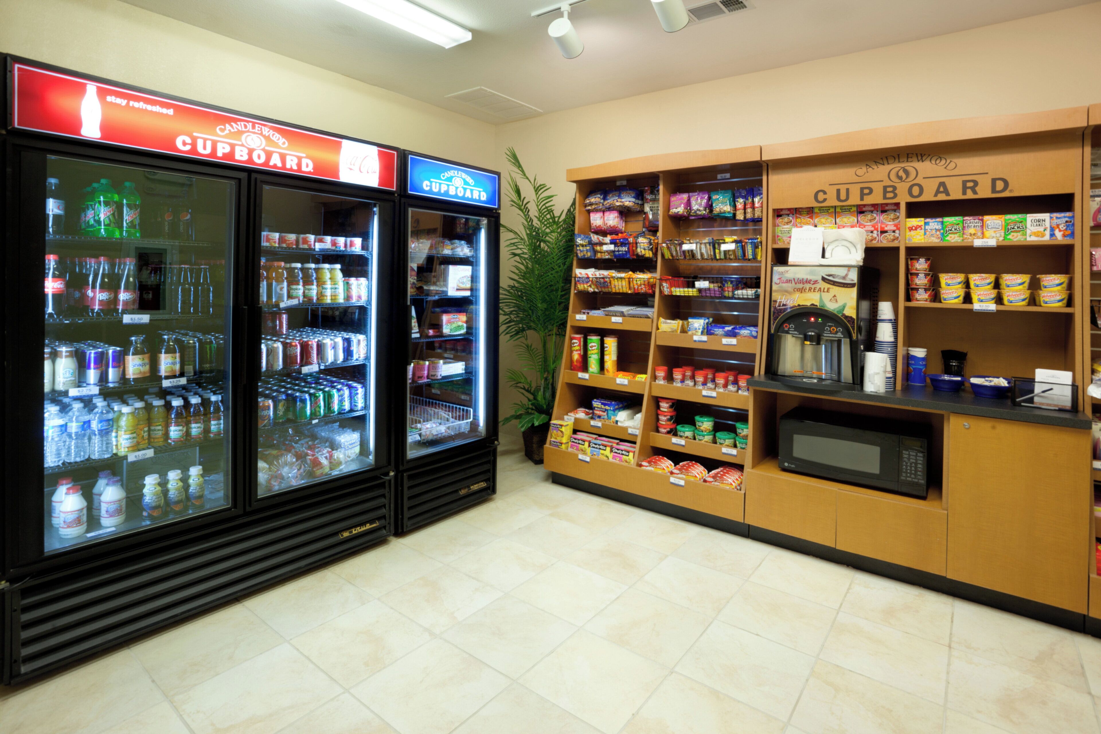 Zona autoservicio de bebidas y alimentos con dispensadores refrigerados, estantes de bocadillos y un microondas.