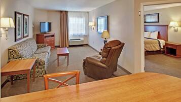 A hotel room with a sofa, armchair, dining table, TV, air conditioning unit, and a bed with a headboard.