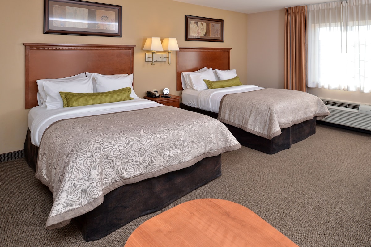 A hotel room with two beds, a nightstand with a lamp, a small table, and a window with curtains.