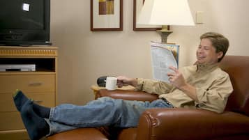 Un hombre sentado en un sillón de cuero, sosteniendo un libro y una taza, con un televisor y una lámpara al fondo.