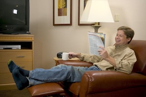 A man sitting in a leather armchair, holding a book and a mug, with a television and a lamp in the background. - Candlewood Suites Williston by IHG (Williston)