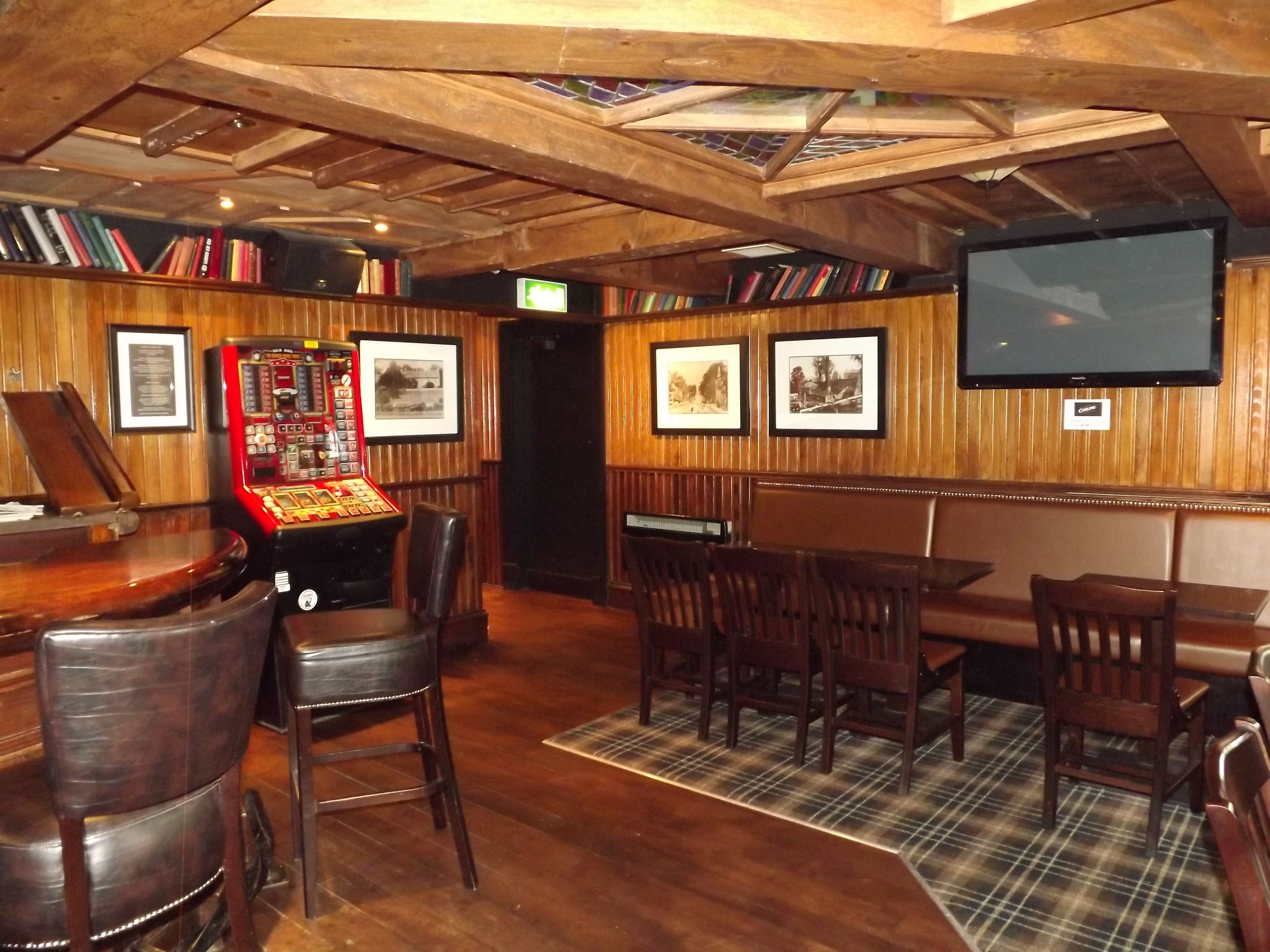 A bar area with wooden ceiling beams, a jukebox, framed pictures on the wall, and a television mounted above a bar counter.