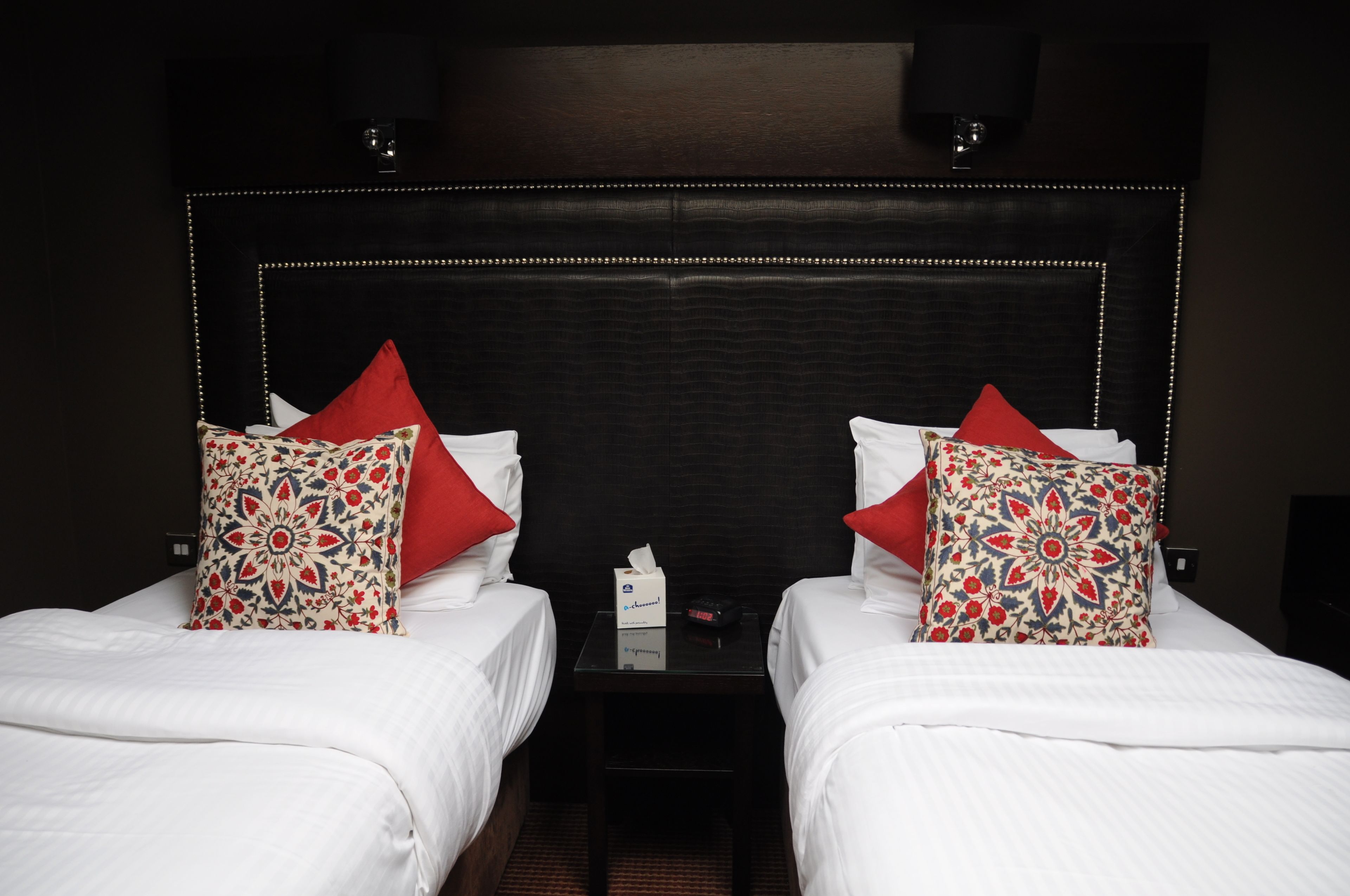 Ein Hotelzimmer mit zwei Betten, jeweils mit weißer Bettwäsche und roten, gemusterten Kissen. Zwischen den Betten steht ein kleiner Tisch mit einer Taschentuchbox und einer Uhr.