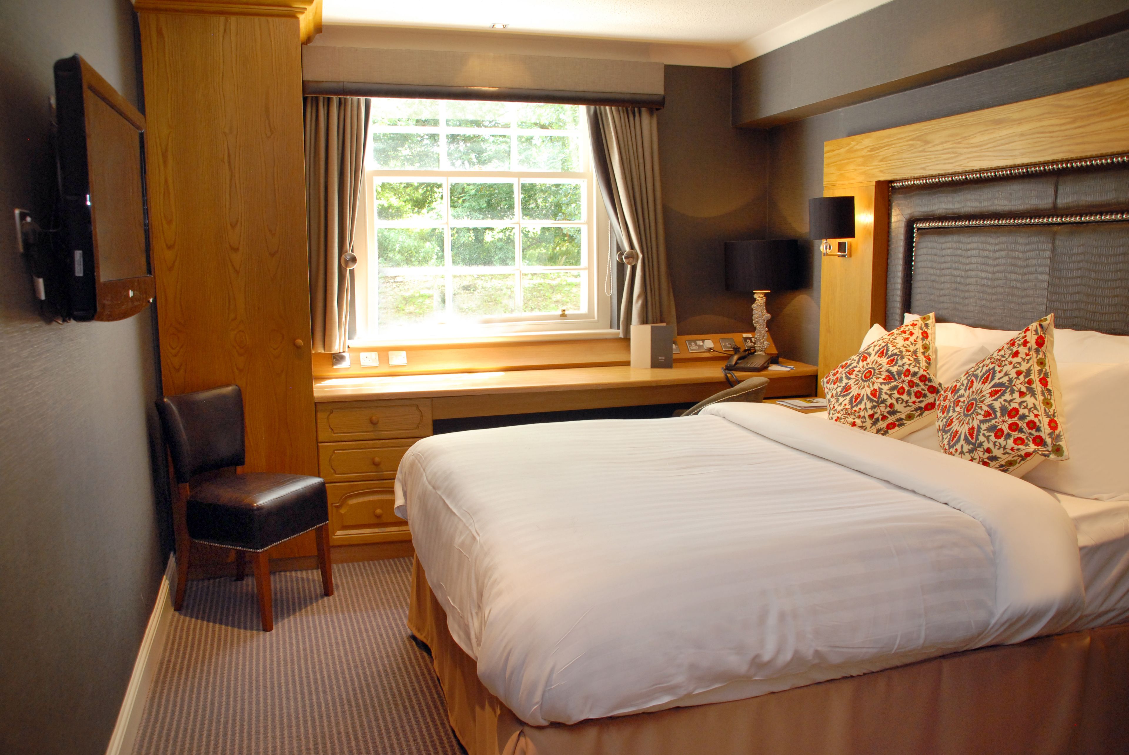 A hotel room with a large bed, a desk with a chair, a window with curtains, and a television on the wall.