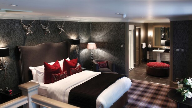 A hotel room with a large bed, a bedside table, a lamp, a red ottoman, a small table with flowers, and a bathroom visible through an open door. - Eglinton Arms Hotel, BW Premier Collection (Glasgow)
