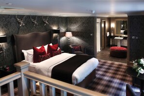 Habitación de hotel con una cama grande, una mesita de noche, una lámpara, un reposapiés rojo, una pequeña mesa con flores y un baño visible a través de una puerta abierta.
