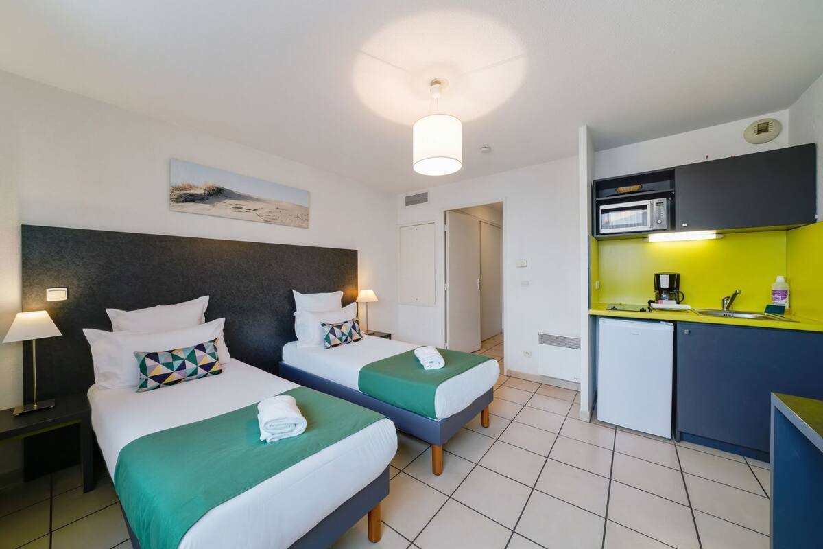 Habitación de hotel con dos camas, kitchenette, una lámpara en el techo y una puerta que da a otra habitación.