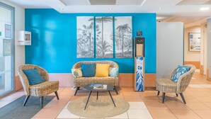 A reception area with a blue wall, a large black and white tropical mural, wicker furniture, and a small round table. - Appart'City Confort La Ciotat Cote Port (La Ciotat)