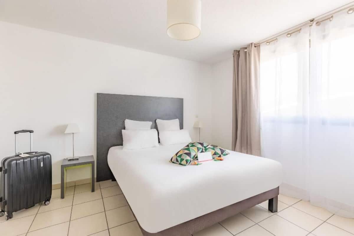 Una habitación de hotel con una cama, una maleta, una mesita de noche y una ventana con cortinas.
