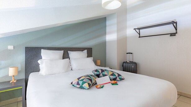 A neatly made bed with white linens and colorful geometric pillows, a bedside lamp, and a suitcase on the floor. - Appart'City Confort La Ciotat Cote Port (La Ciotat)